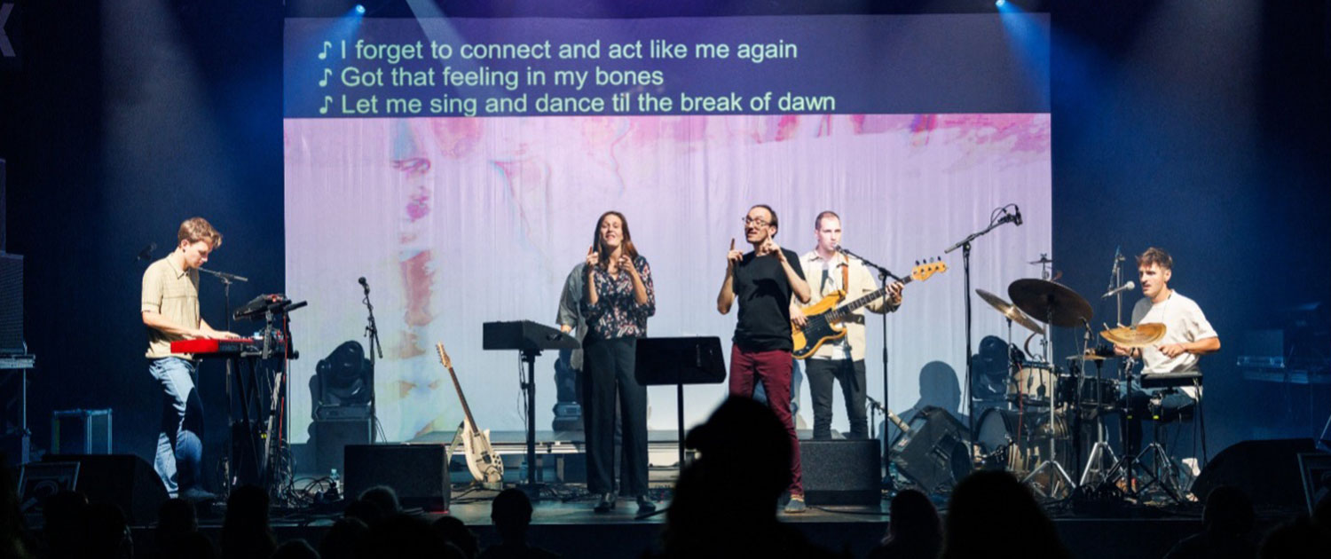 Auf einer Bühne sind fünf Personen zu sehen. Sie stehen vor einer Leinwand. Auf der Leinwand gibt es oben drei Zeilen mit dem englischen Liedtext, der gesungen wird. Dieser Text wird von Schriftdolmetschern produziert. Die zwei Personen in der Mitte der Bühne sind Gebärdensprach-Poet:innen. Sie inszenieren den Text in Gebärdensprache. © Beat the Silence mit Link: Das WUK | Beat the Silence