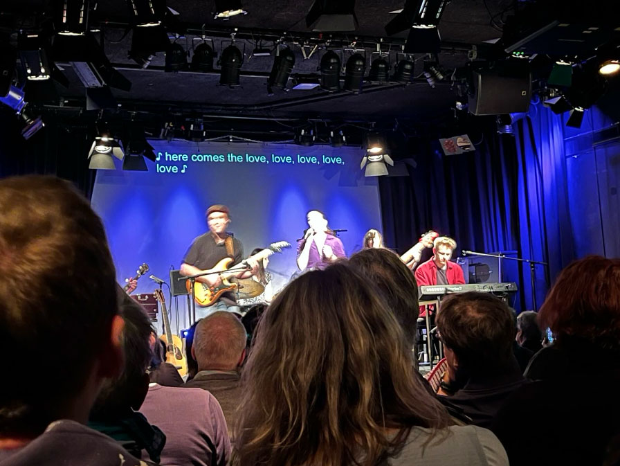 Auf einer Bühne sind im Hintergrund drei Personen zu sehen. Sie singen und spielen Musikinstrumente. Im Vordergrund sind die Konzertbesucher:innen. Sie sehen die Musiker:innen auf der Bühne und die Live-Übertitel in englischer Sprache. © Beat the Silence mit Link: Das WUK | Beat the Silence