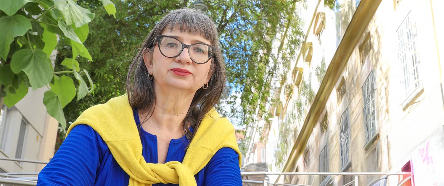 Eine Frau mittleren Alters mit markanter Brille und rotem Lippenstift blickt sitzend in die Kamera. Sie trägt eine blaue Bluse, darüber locker über die Schultern gebunden einen gelben Pullover. Hinter ihr ein Baum und eine Häuserzeile. Es ist Ursula Nasswetter.