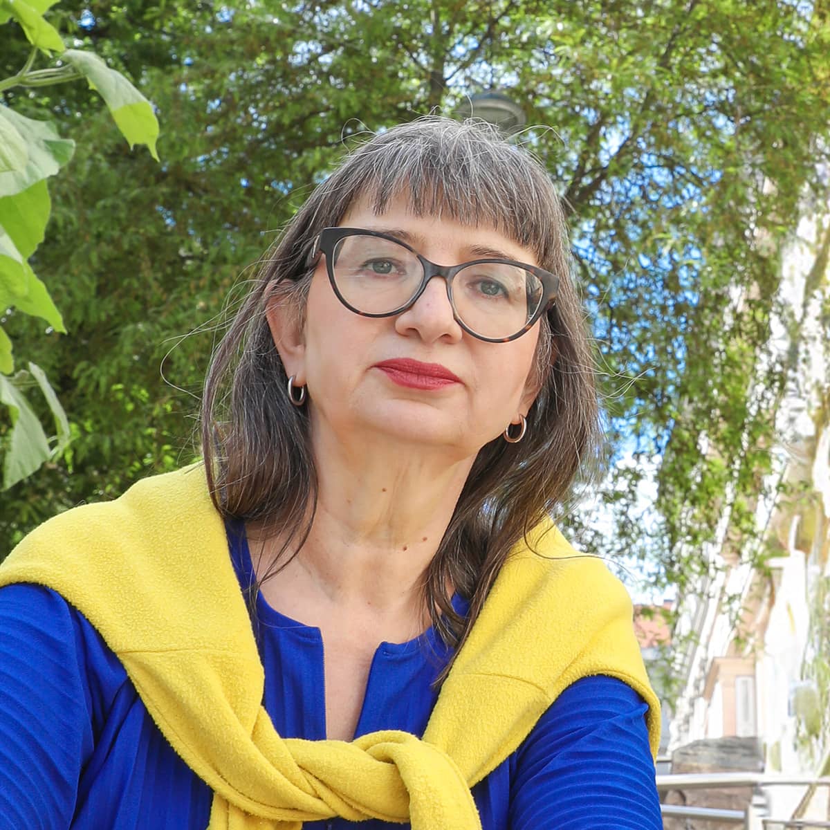 Eine Frau mittleren Alters mit markanter Brille und rotem Lippenstift blickt sitzend in die Kamera. Sie trägt eine blaue Bluse, darüber locker über die Schultern gebunden einen gelben Pullover. Hinter ihr ein Baum und eine Häuserzeile. Es ist Ursula Nasswetter.