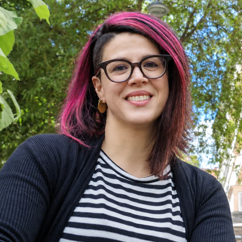Barrierefreie Kommunikation: Erfolgreiche Kommunikation durch Expertise und Weitblick Eine Frau mit rosa-schwarzen Haaren und Seitenscheitel, brauner runder Brille und goldenen Ohrringen lächelt in die Kamera: Es ist Verena Brinda